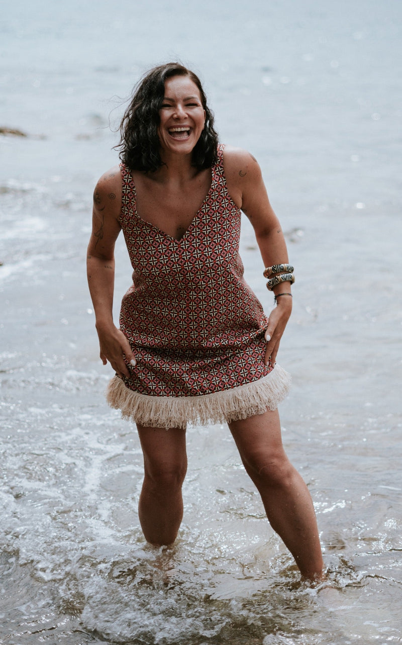 A woman stands in shallow ocean water wearing a sleeveless rust-brown patterned dress with a cream fringe hem and stacked bracelets on one arm, with a shoreline and trees in the background.